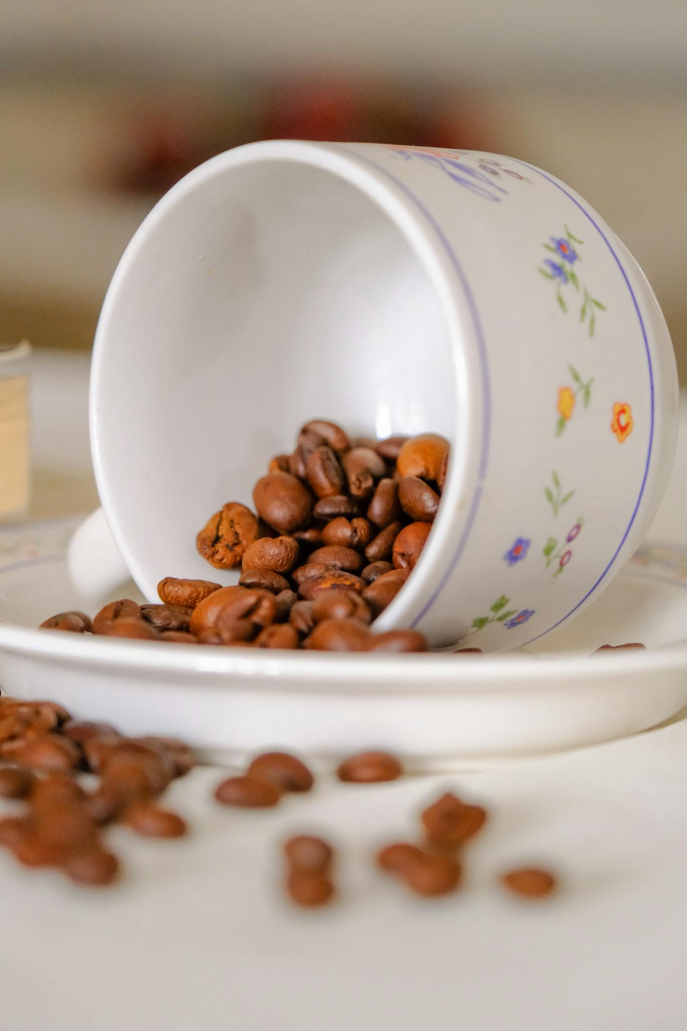 Floral mug tipped over with aromatic coffee beans spilling onto a white saucer.